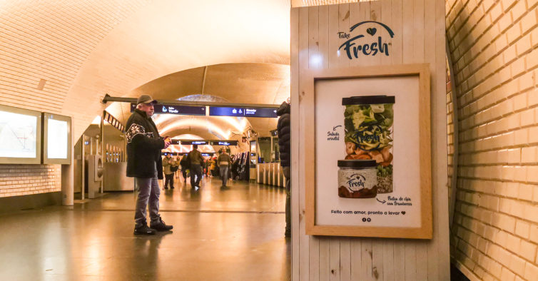Homem olhando para a vending machine da empresa Take Fresh em estação de metrô. Ao fundo pessoas transitam no corredor